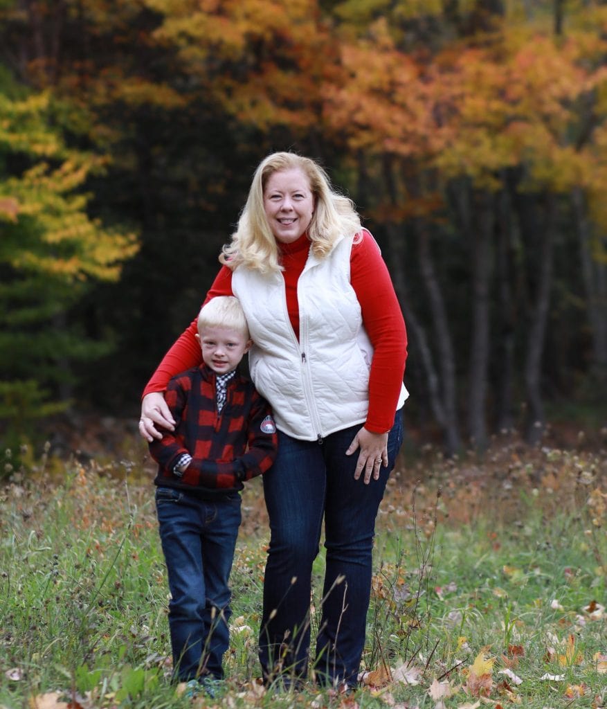 mothers arm around son, standing in open field with fall trees in the background