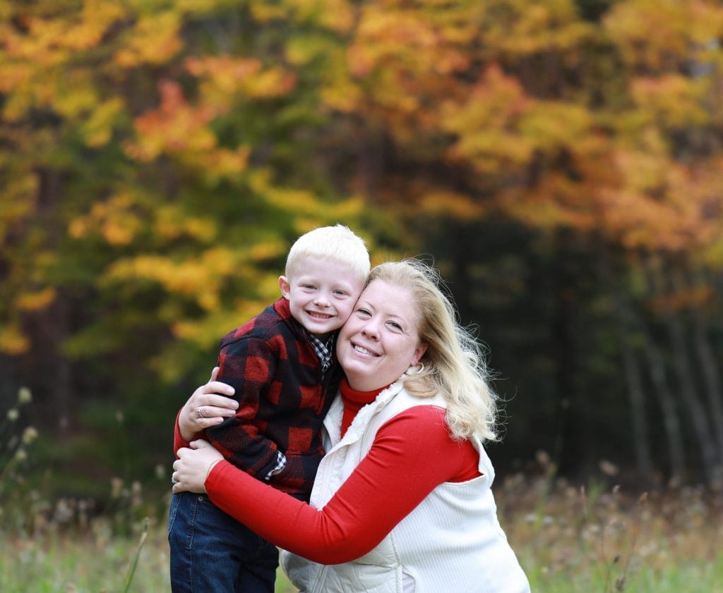 mom bent down hugging her son, fall trees in the background