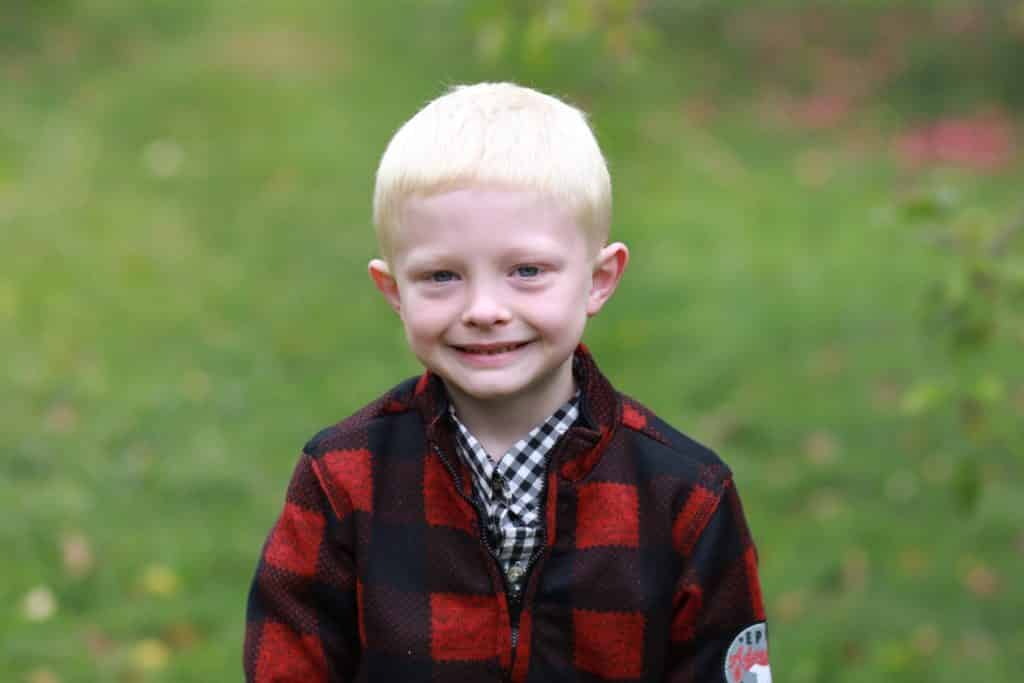 boy in plaid jacket standing in apple orchard starring at the camera with a smile.