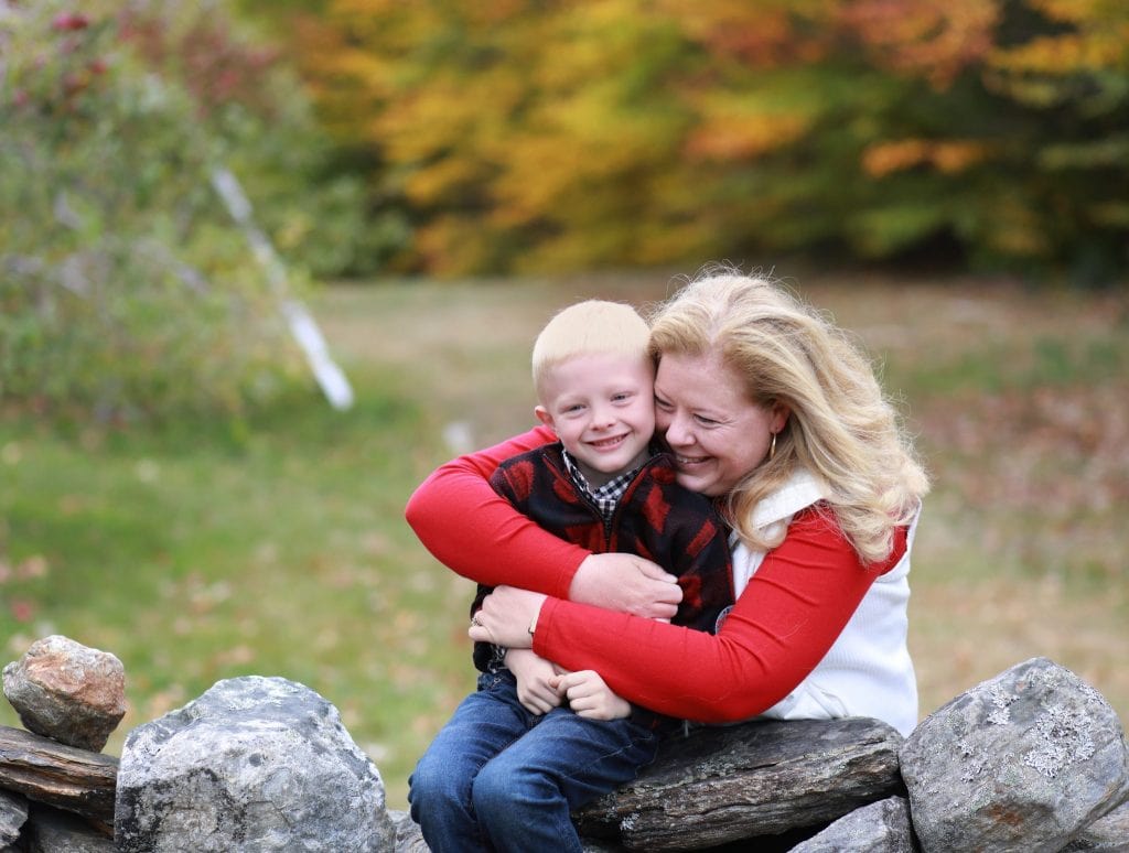 mom looking at son hugging her son from behind rock wall, fall trees in the background