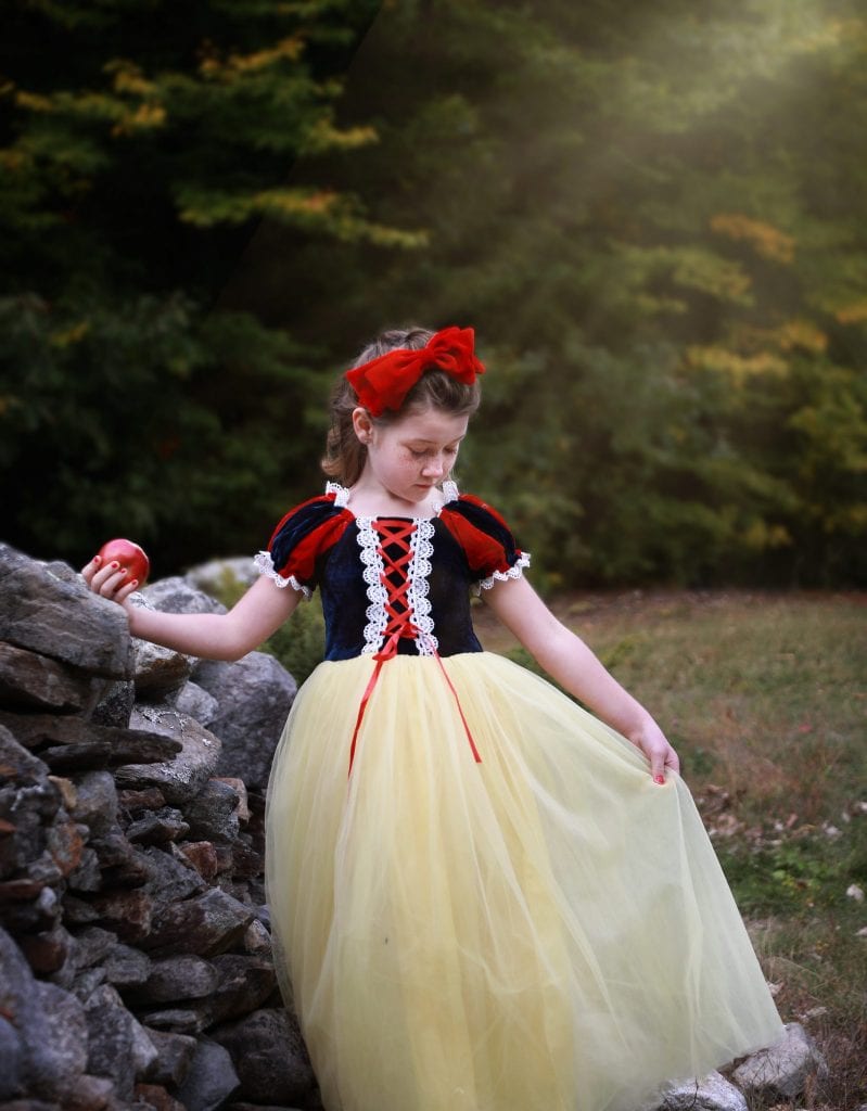 Little girl dressed up as snow white leaning on a grey stone wall holding out her dress looking down, apple in hand
