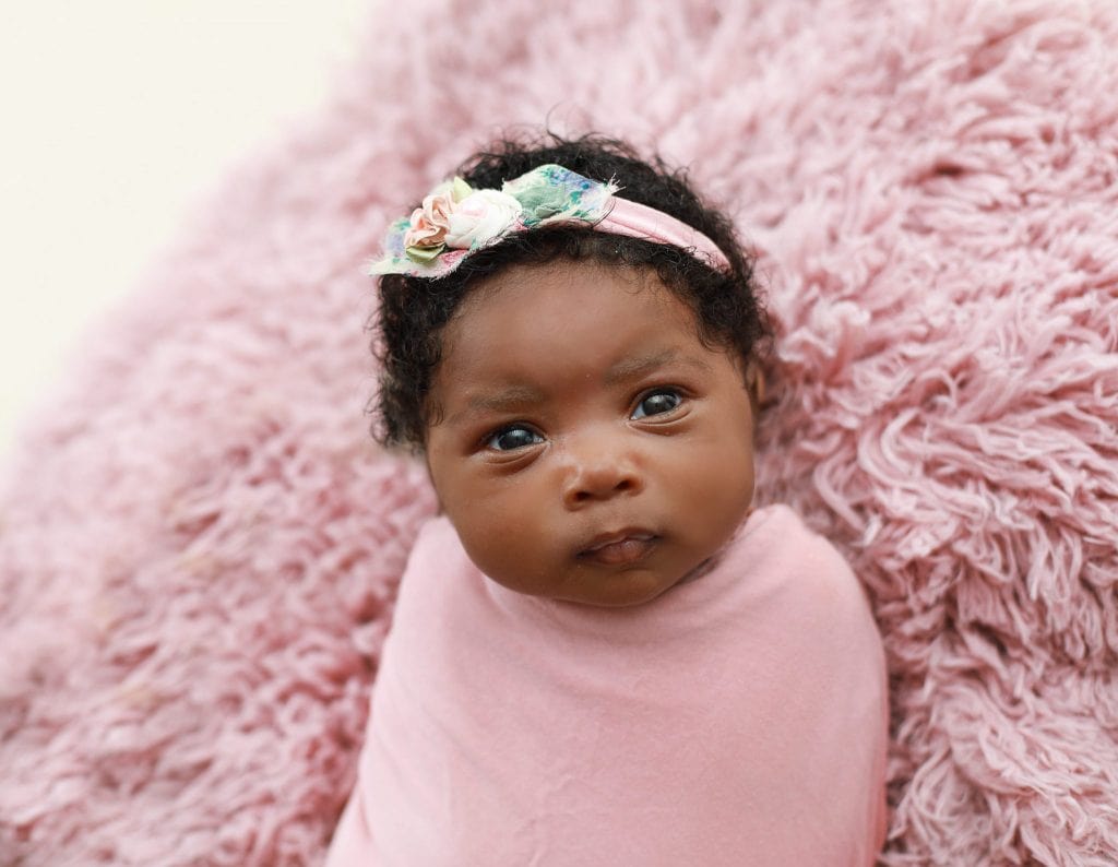 five week old newborn girl wrapped in pink laying on pink fur with a pink floar headband, looking at the camera
