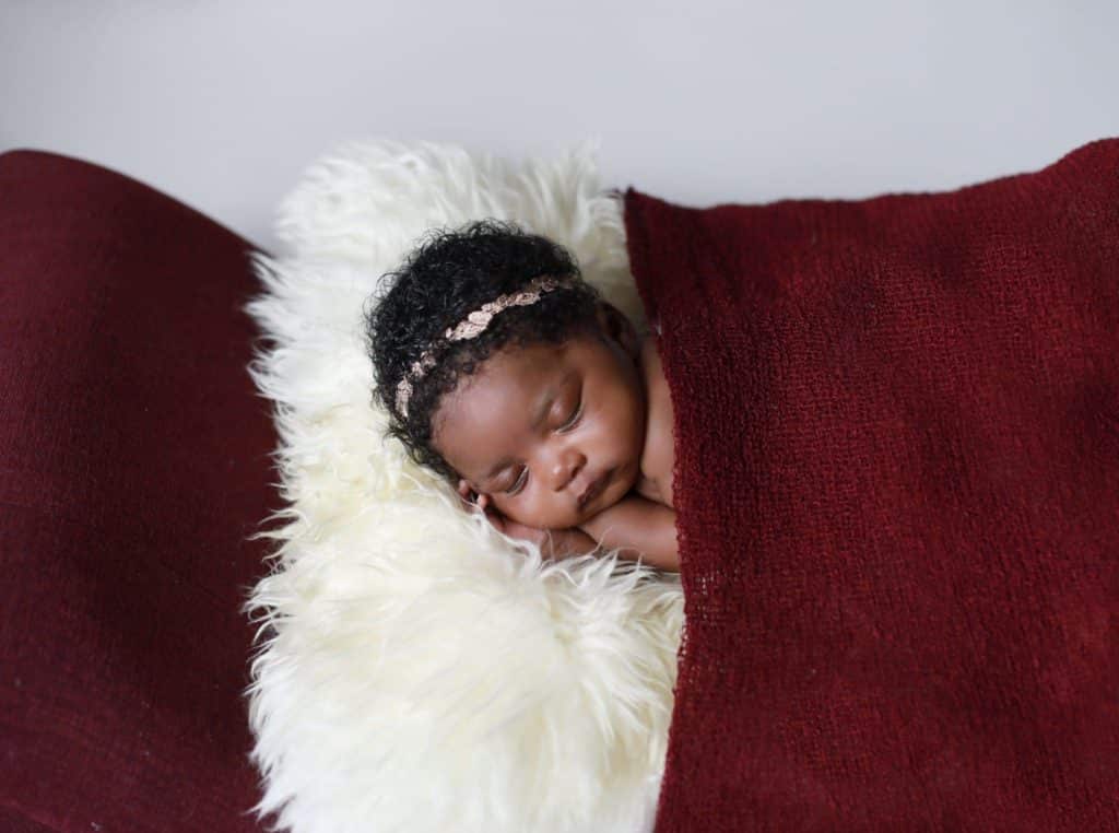 dark skinned newborn girl laying on stomach on red chase lounge, white fur, covered with a red wrap like a blanket, neutral headband