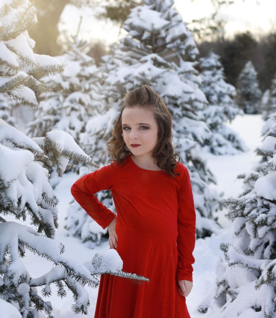 Young girl red dress, curly hair, hair half up and half down, red dress, hand on her hip, looking at the camera, tree farm, snow covered trees, smiling