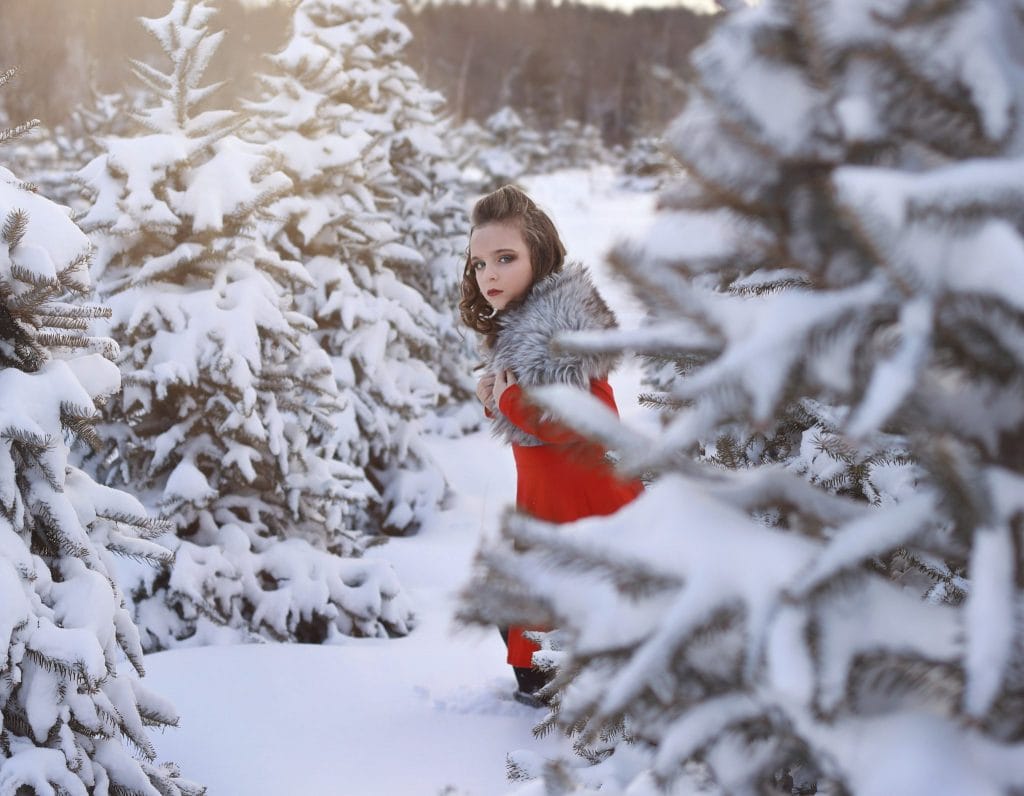 young girl running, curly hair, red dress, grey faux fur, looking sideways to the camera, tree farm, snow covered trees, winter, curly hair half up and half down