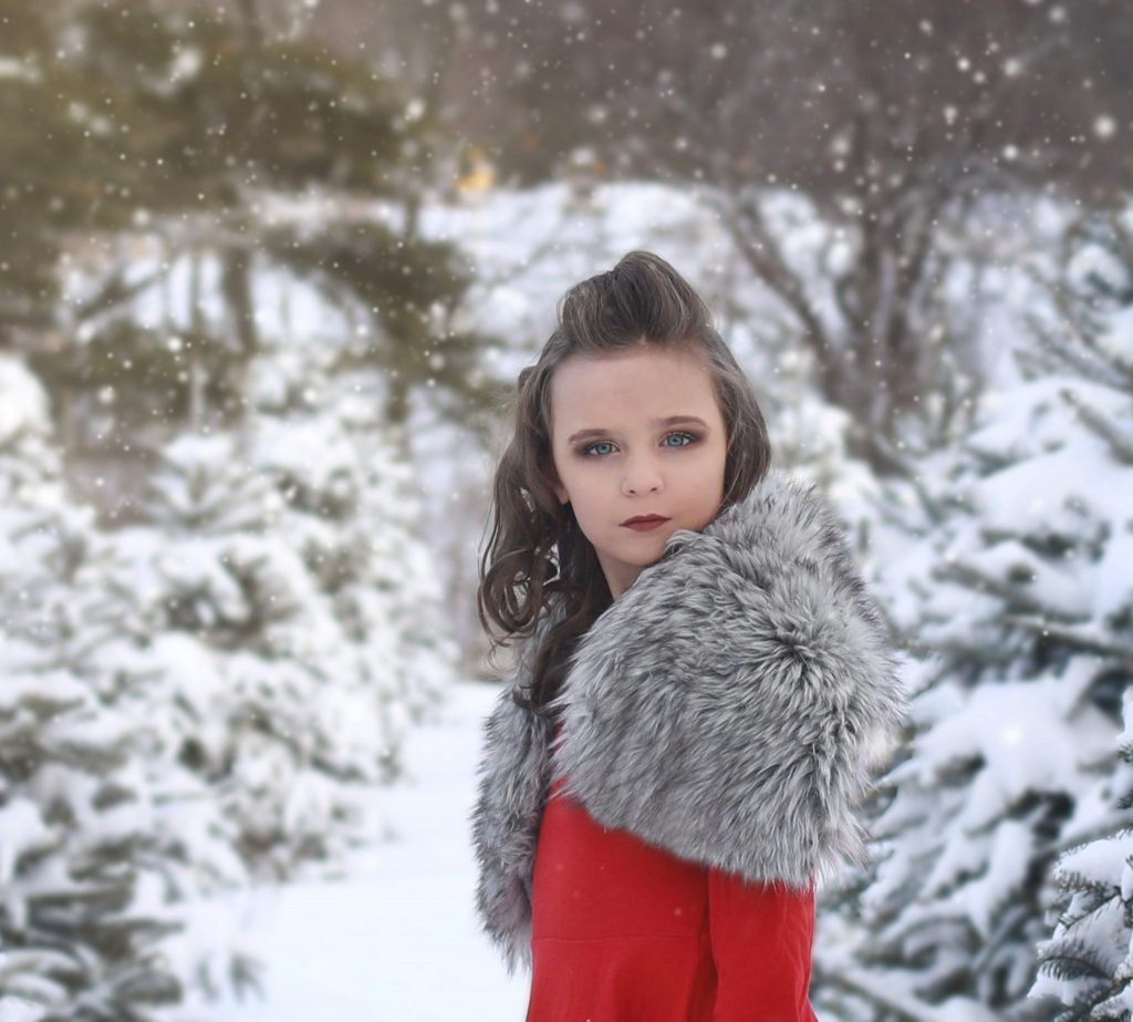 young girl, curly hair, red dress, standing sideways, looking at the camera, tree farm, snow covered trees, snowing