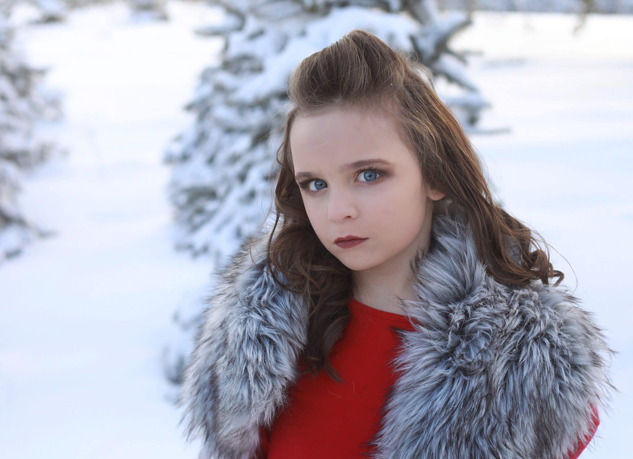 young girl, small smirk, red dress, tree farm covered in white snow, grey faux fur on shoulders