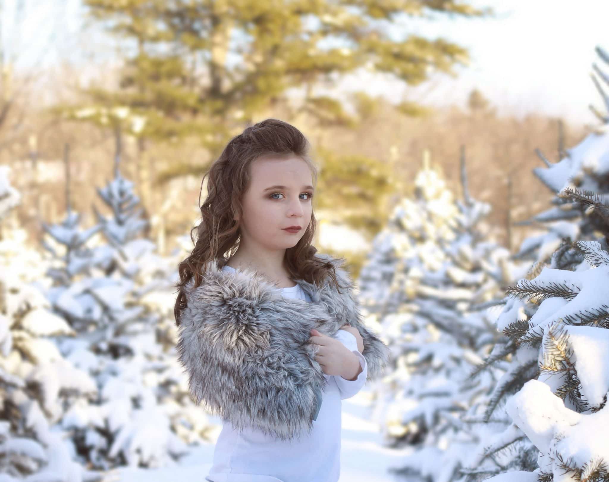 Young girl, white shirt, grey faux fur on her shoulders, curly hair, tree farm, snow covered trees, winter, sideways looking at the camera
