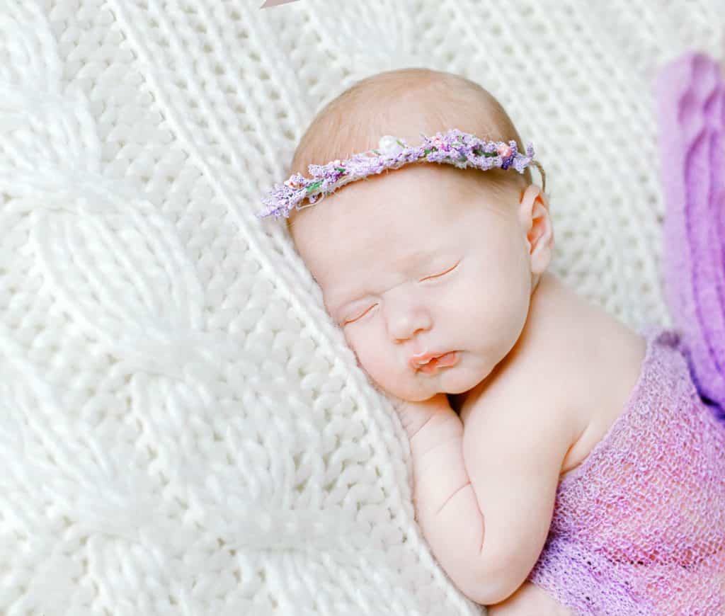 Newborn baby girl laying on her stomach wrapped in lavender with ehr hands under her chin facing the camera