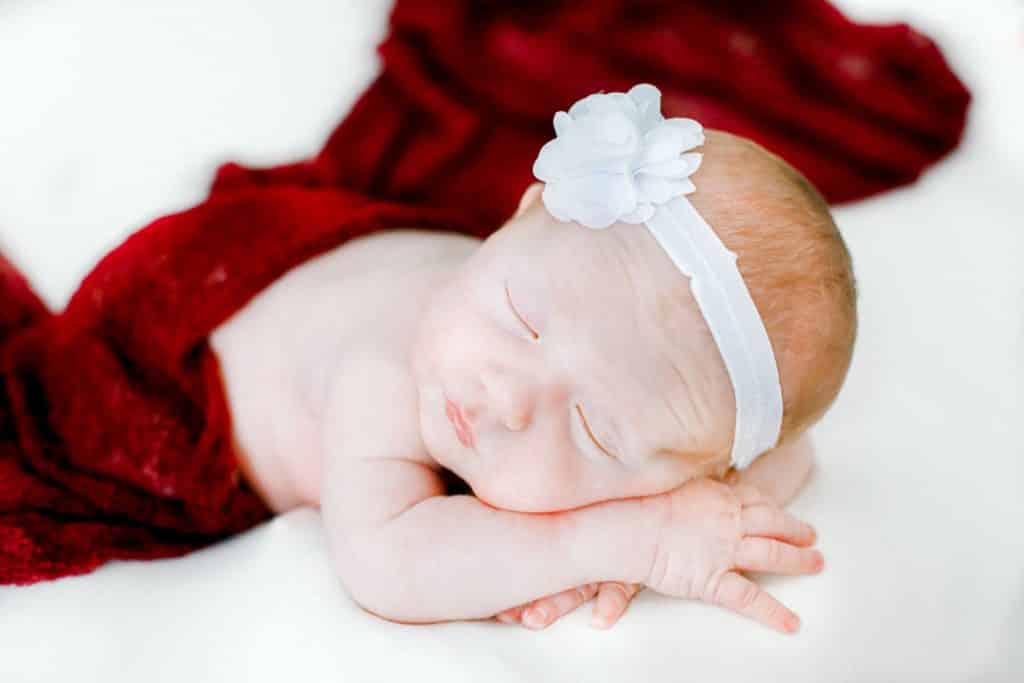 newborn baby girls laying on her stomach with a maroon cover up and a white headband on a white blanket