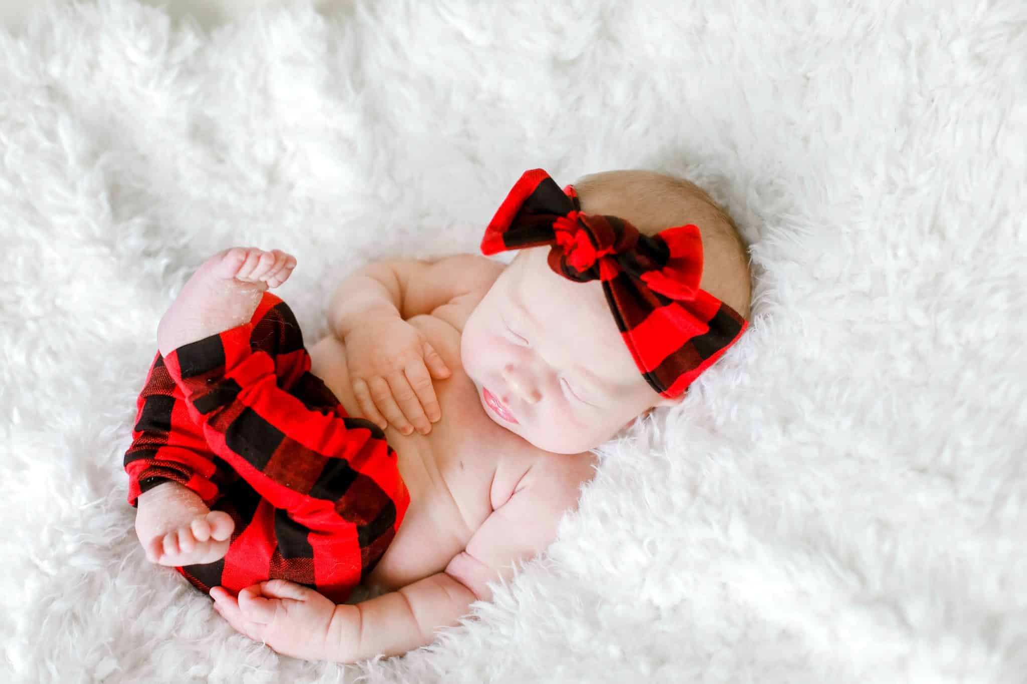 newborn baby laying on her back in red plaid pants and a bow with ehr feet curled up on a white blanket