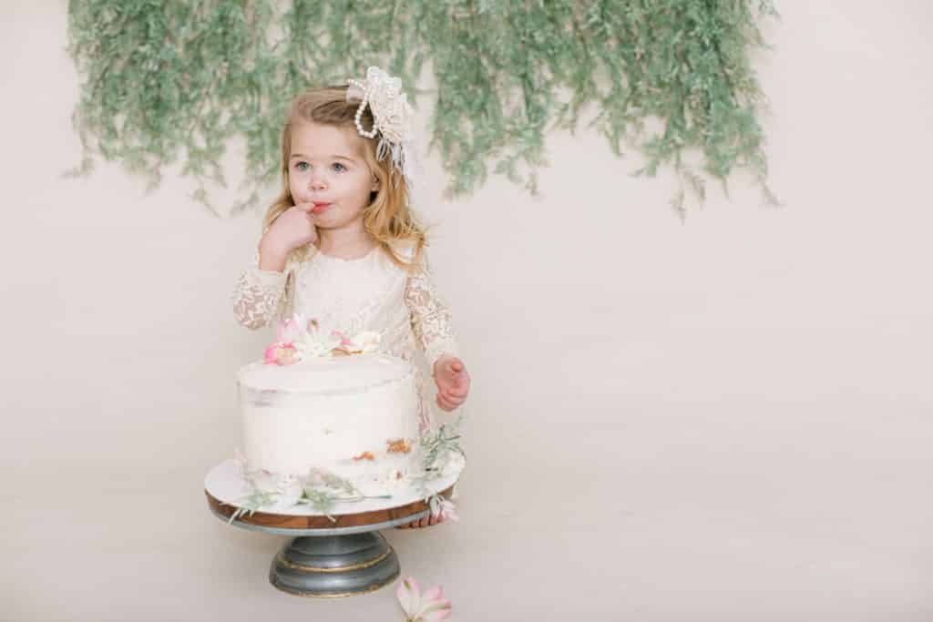 one year old girl in white dress looking at the camera eating cake off her finger