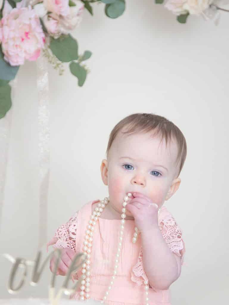 One year girl in pink dress looking at the camera sucking on pearls