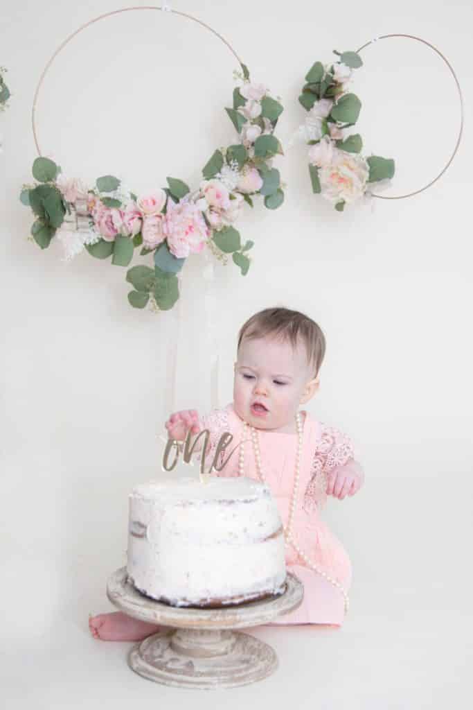 one year old girl sitting in front of her cake looking at the cake with her hand on it