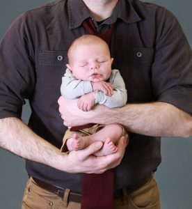 dad holding newborn, hand on feet and other hand on waist, newborn pressed against his chest facing the camera