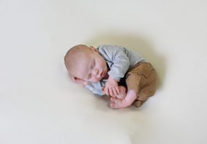 newborn boy curled up in tan pants and grey long sleeved, grabbing foot, hand on ear, laying on his side.
