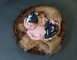 newborn baby curled up in brown nest, burlap, navy blue deer pants and hat