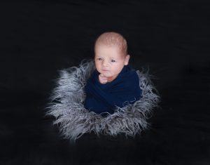 newborn wrapped in blue wrap upright on grey fur awake