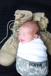 newborn wrapped in white in military hat with military boots on black backgroun