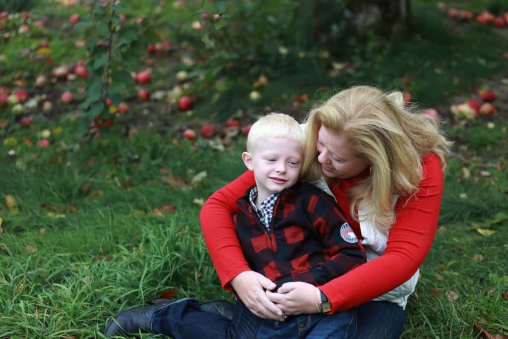 mom and son sitting under an apple tree with apples around them, mom hugging son from behind looking at him smiling.