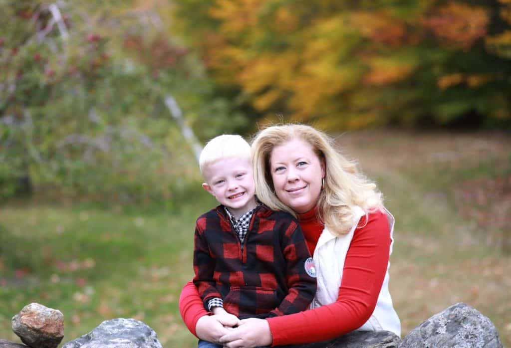 mom hugging son from behind rock wall, fall trees in the background