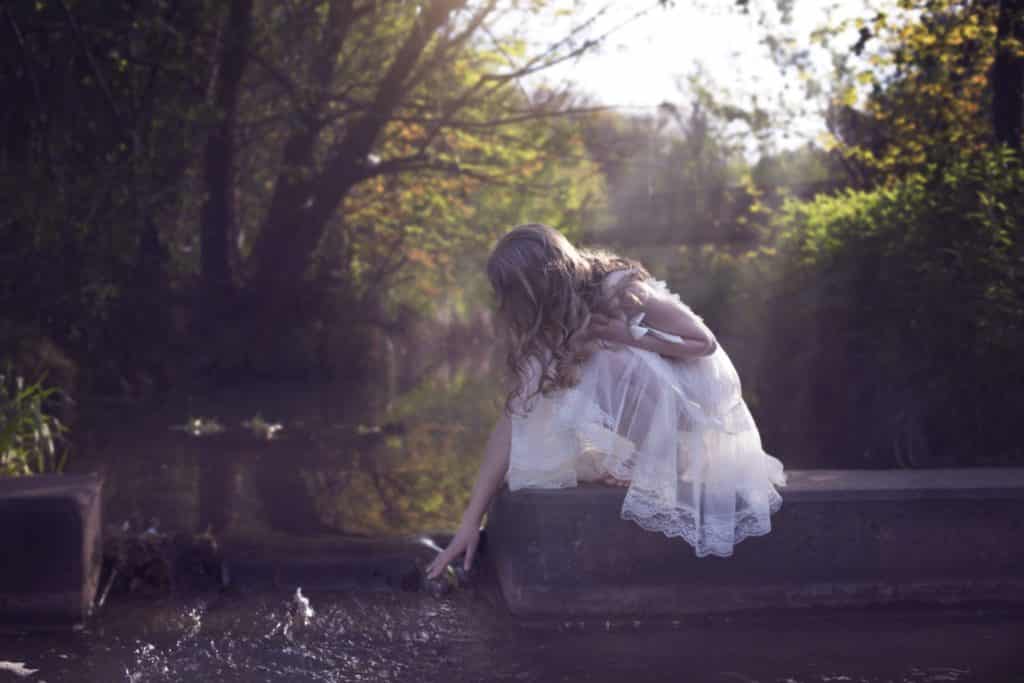 young white girl dressed in white vintage dress reaching down and touching the water facing away from the camera for fairy tale photography