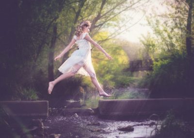 young white girl dressed in white vintage dress jumping over water with sun coming in from the right