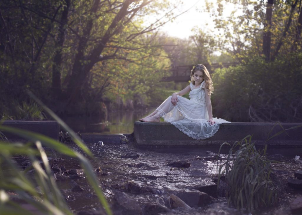 Young white girl sitting on stone over water looking into the water with sun coming through the right side fairy tale photography