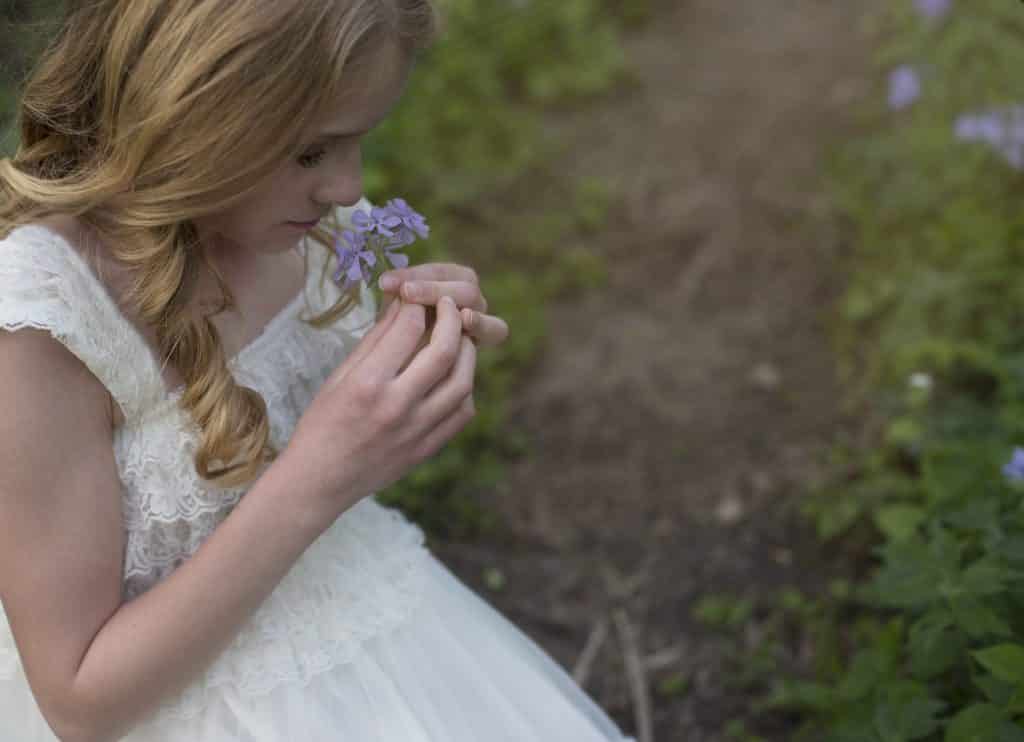 young white girl bending down in tthe forest smelling a purple flower, sun coming in from the right