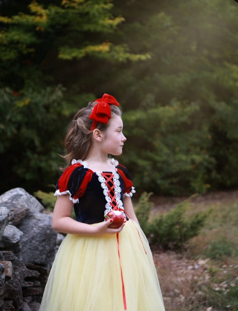 little girl dressed as snow white holding an apple near grey stone wall looking out to the pple orchard, light shining down on her