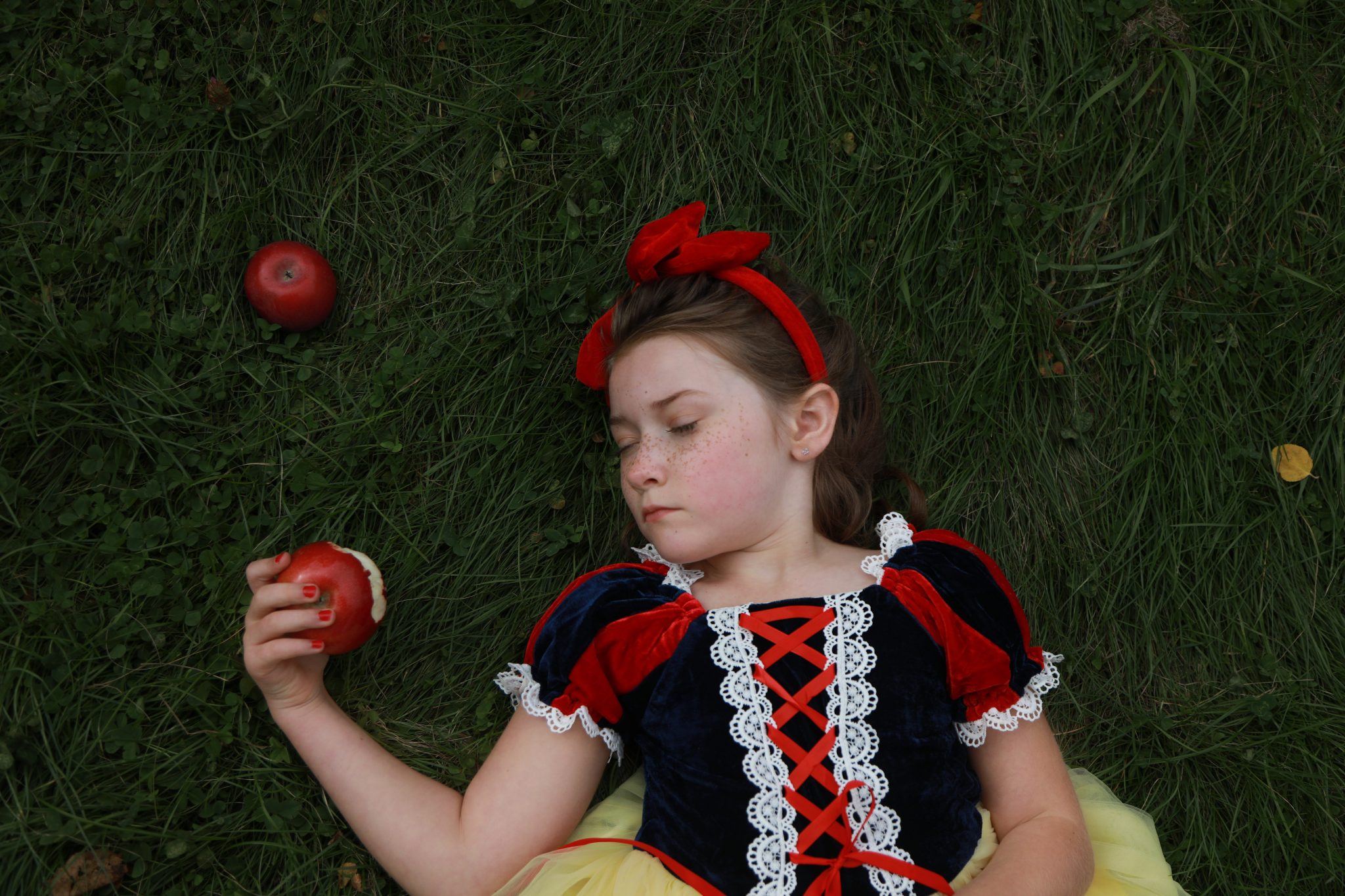 little girl laying on grass dressed up as snow white, holding a bitten apple with her eyes closed.