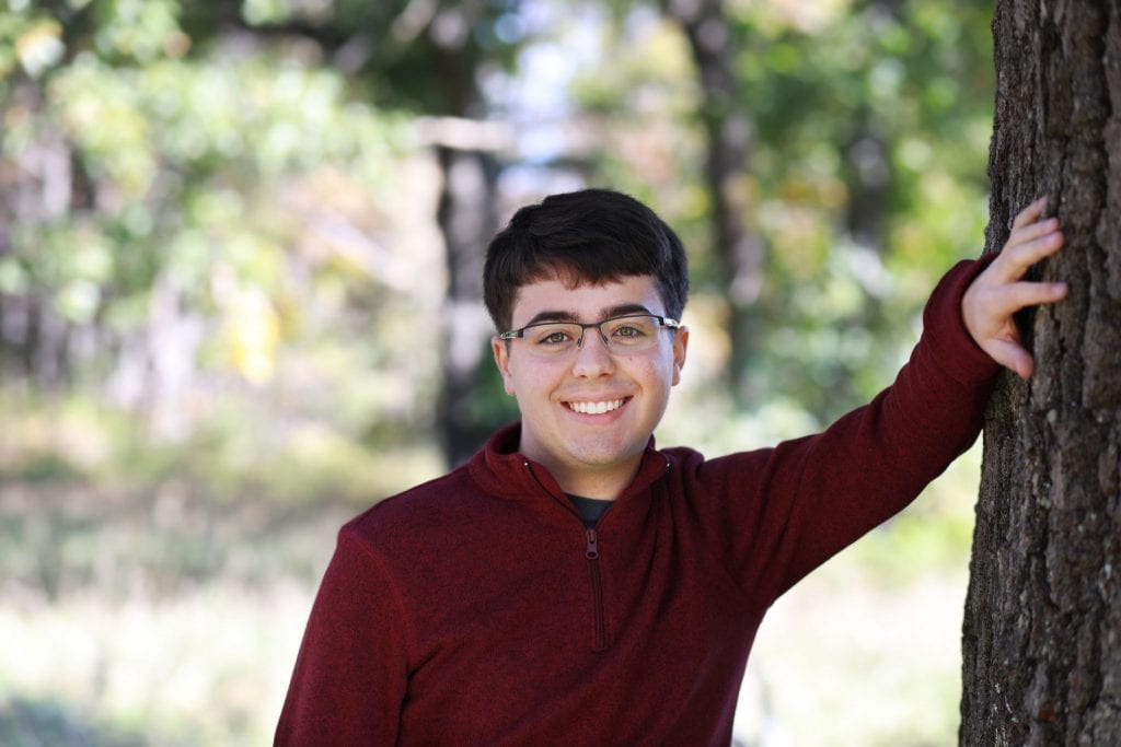 senior boy leaning against a tree with one arm up, the other at his side, wearing glasses and a red shirt, smiling, teeth showing