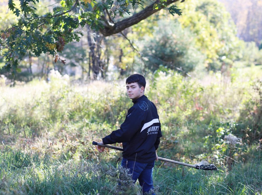 senior boy wearing custome lacrosse jacket, standing in an open field, back facing the camera, turning his head to the camera holding his lacrosse stick