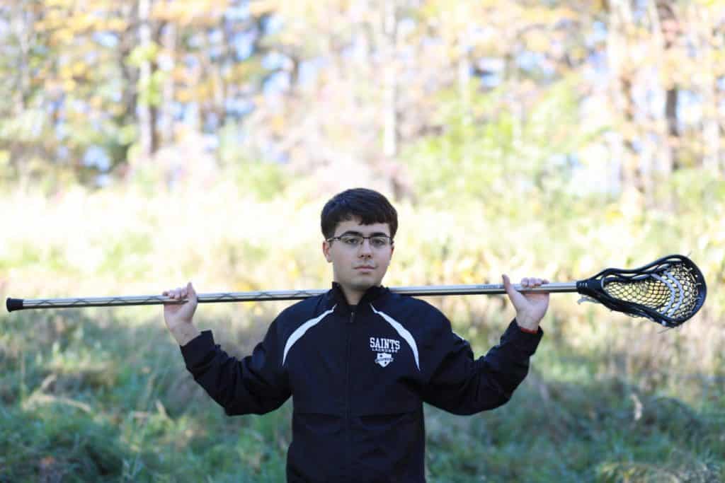 senior boy standing in a field holding his lacrosse stick on his shoulders wearing his lacrosse jacket with his glasses