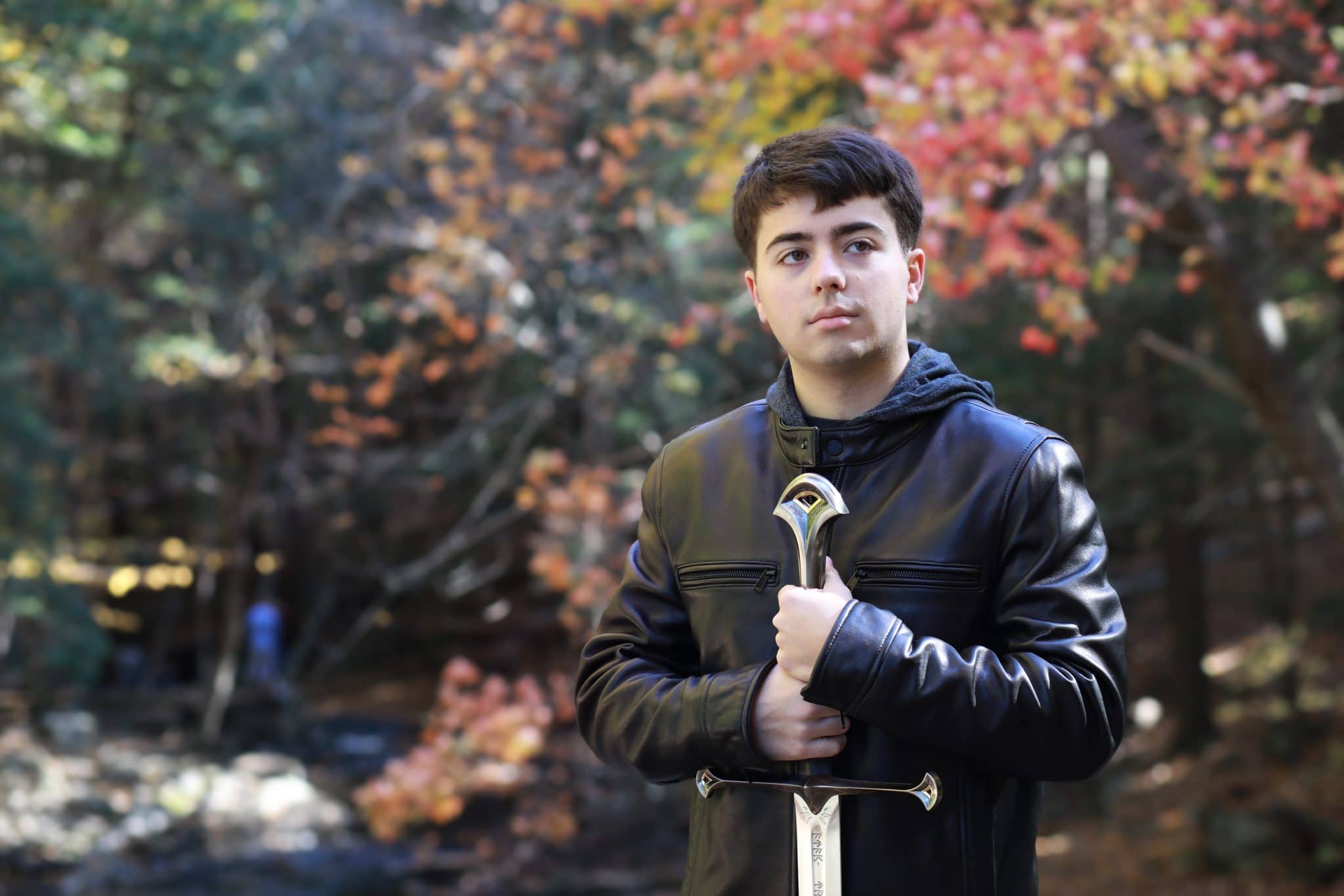 Senior boy in leather jacket holding a giant sword on a bridge with fall colored trees in the background