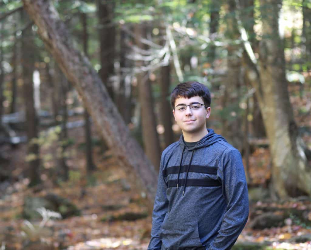 Young man senior in High School in the woods, hands in pocket, wearing glasses, surronded by fall colors on the trees, standing sideways taking his senior picture