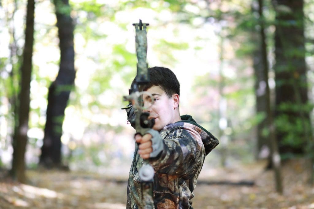 senior boy in camouflage jacket, holding bow and arrow, looking at the camera like he would shoot the bow and arrow in the woods.