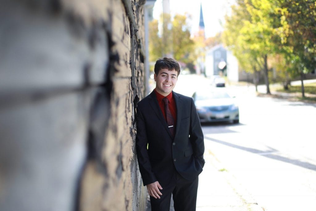 senior boy leaning on brown and black textured building in a black and red suit with a red tie, wearing glasses, urban style, hand in his pocket and one hand out