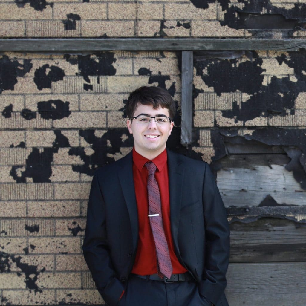 senior boy standing against textured building, black and brown in a black suit with a red shirt and red tie, wearing glasses