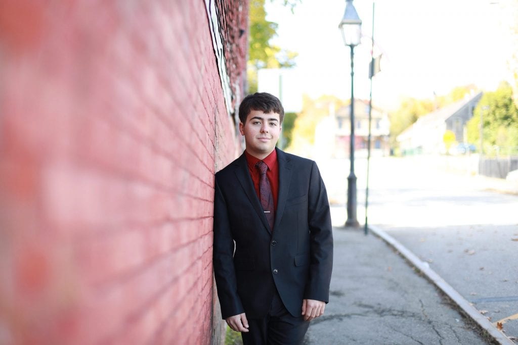 senior boy leaning against red brick building sideways, hands hanging at his waist, black and red suit 