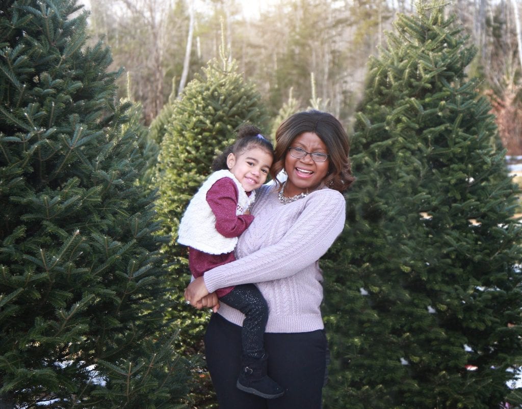 mom holding two year old daughter standing between green trees in the tree farm