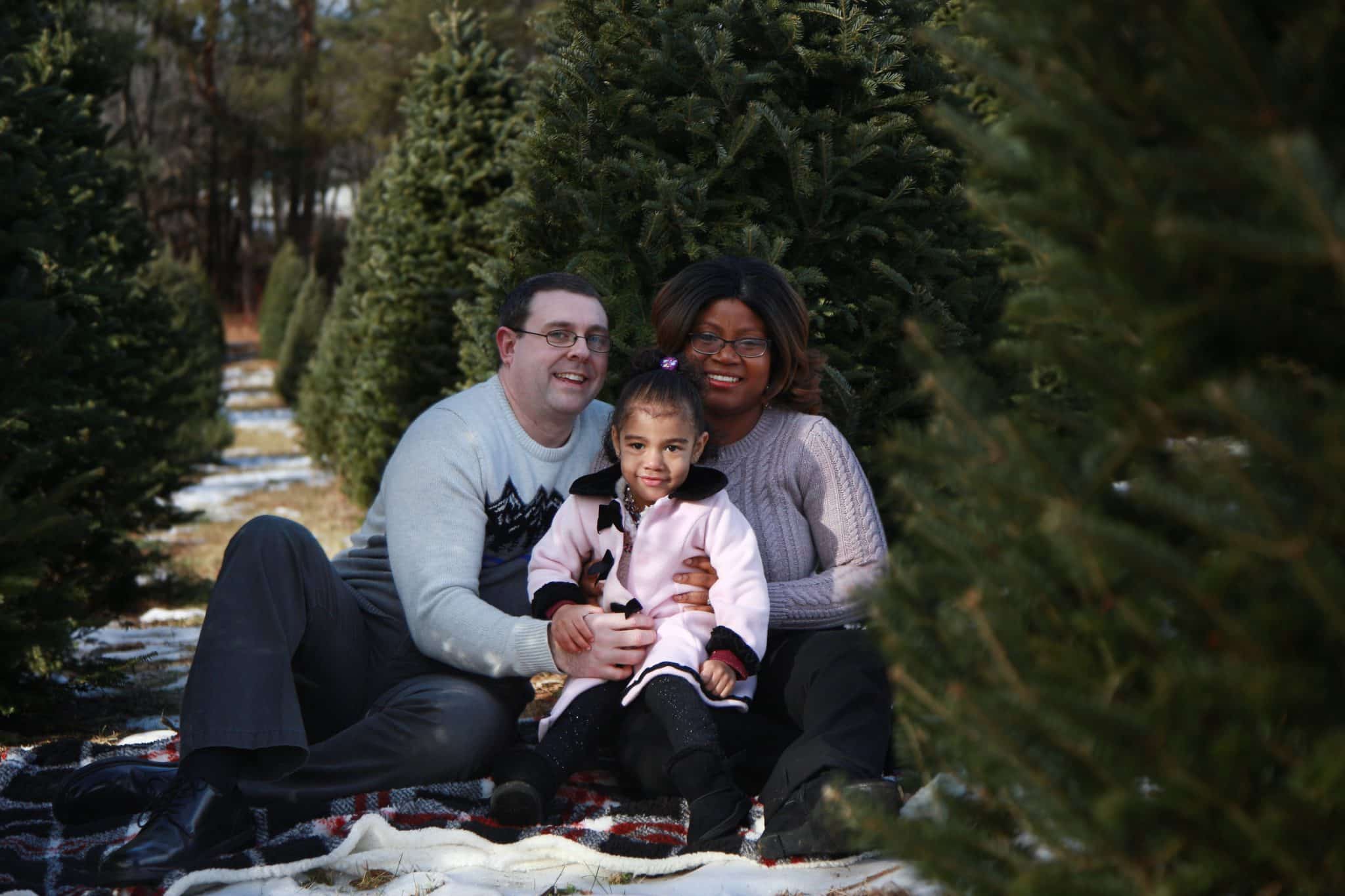 mom, dad, and two year old daughter sitting on a white blanket at a tree farm surrounded by trees hugging each other