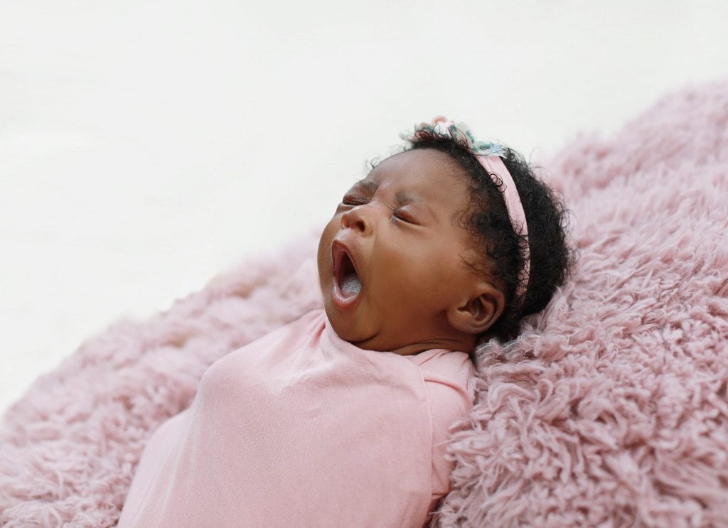 newborn girl laying on pink fur, yawning,wrapped in a pink wrap, pink floral headband 