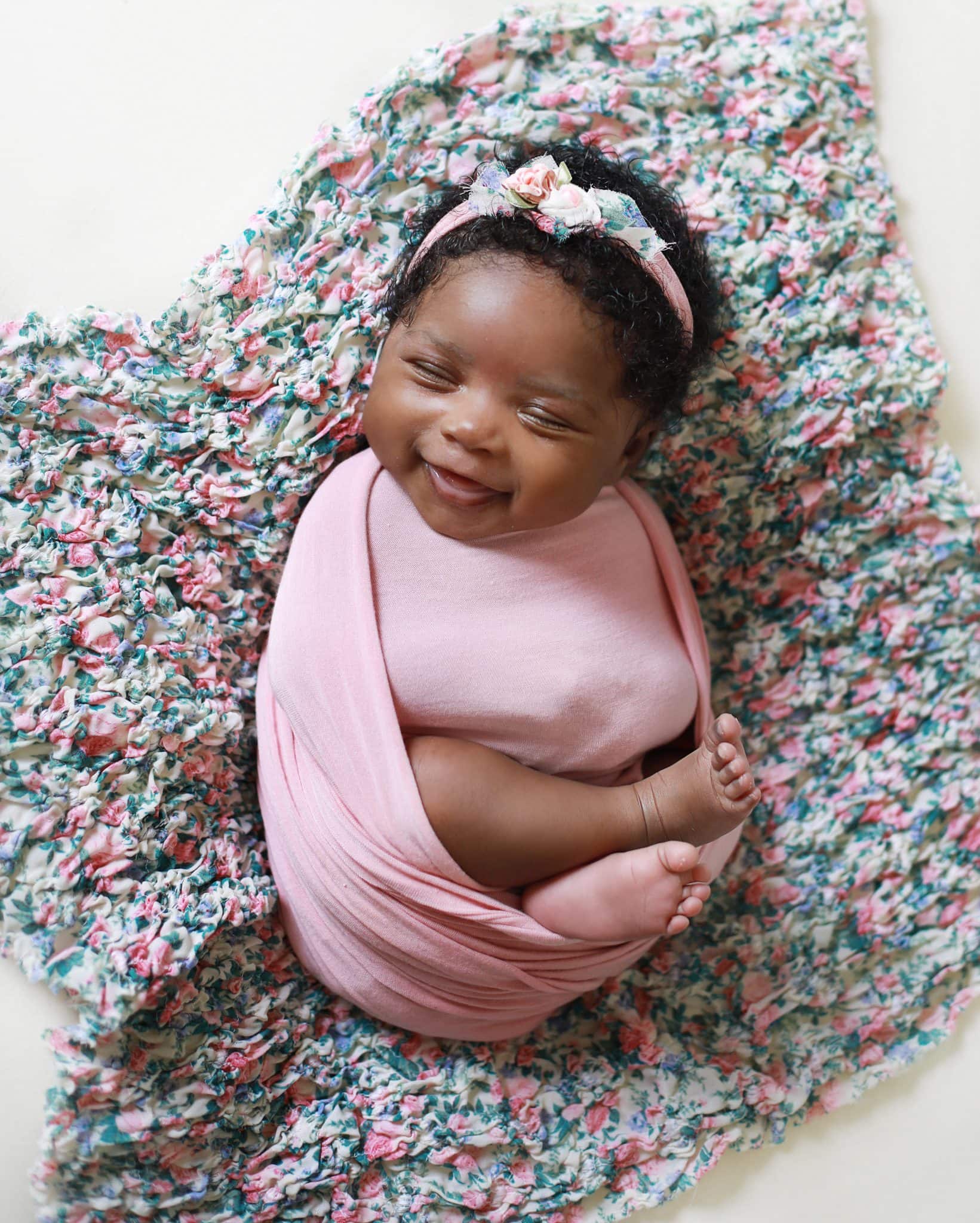 newborn girl, dark skin, wrapped in pink on a floralprint background smiling