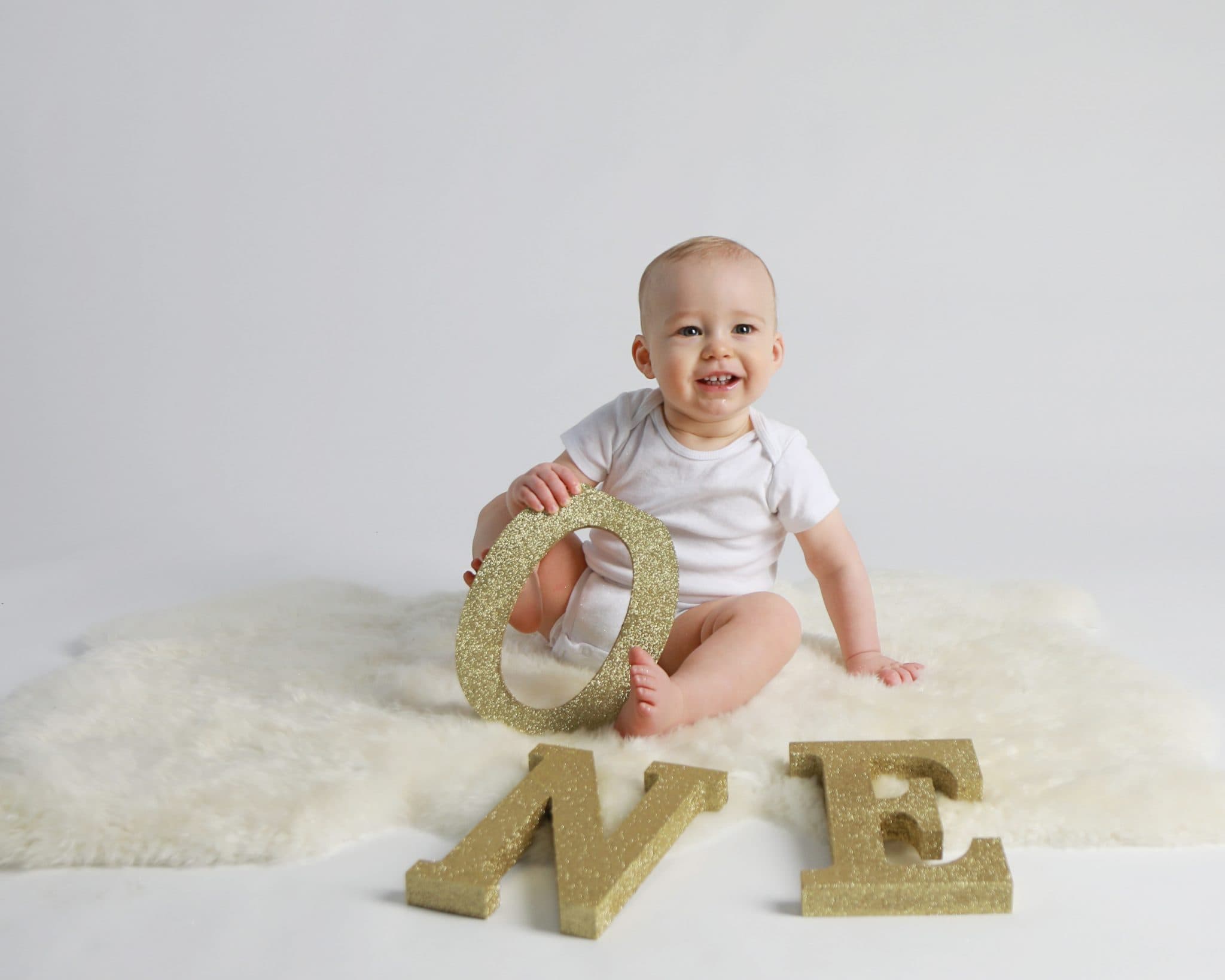 Baby Boy in white onsie with gold letter s that spell one, on a white back ground andsitting on white fur holding the O