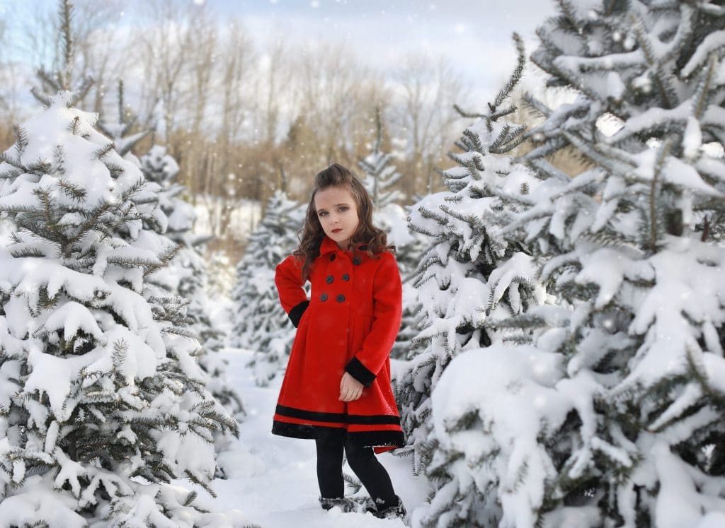 young girl, red coat, looking at the camera, hand on her hip, curly hair, black boots, tree farm, snow covered trees, winter, 