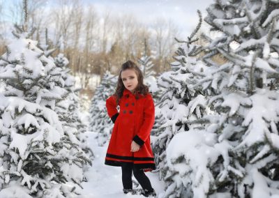 young girl, red coat, looking at the camera, hand on her hip, curly hair, black boots, tree farm, snow covered trees, winter,