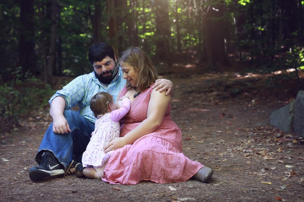 mom in pink dress nursing baby in pink dress, dad in jeans arm around wife in nature