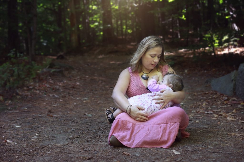 Mom in a pick dress sitting down nursing baby in a pink dress, on a wood path sun coming in above them