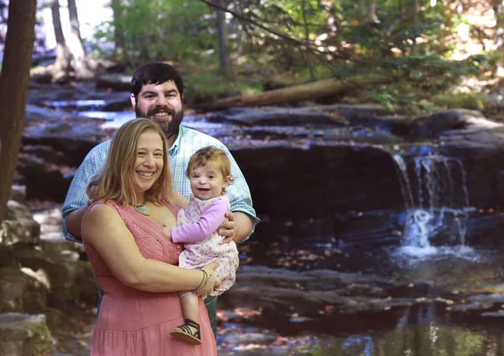 mother and father and toddler in woods, pick dress and jeans, sun peeking in 